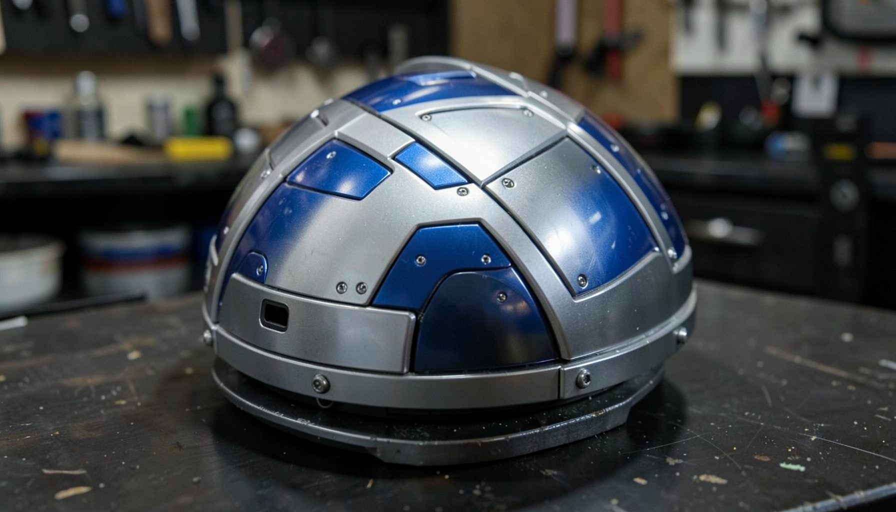 A polished aluminum astromech dome with blue and silver panel details resting on a dark workshop surface
